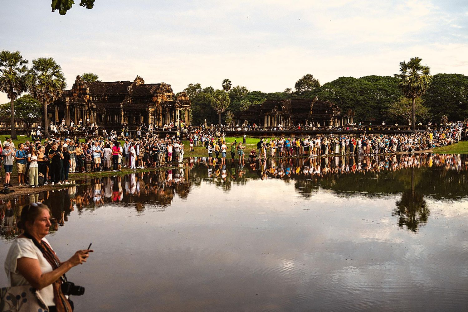 A large crowd gathers at a lakeside park during a summer event with reflections visible in the calm water.