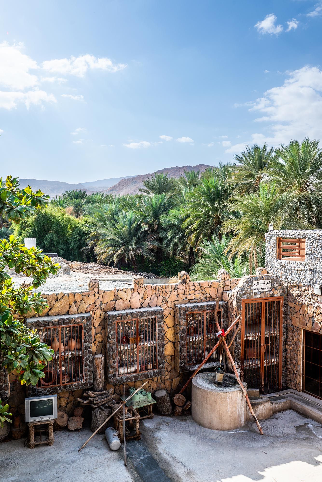Traditional stone building with rustic gates and palm trees in a desert oasis setting against mountain backdrop and blue sky.