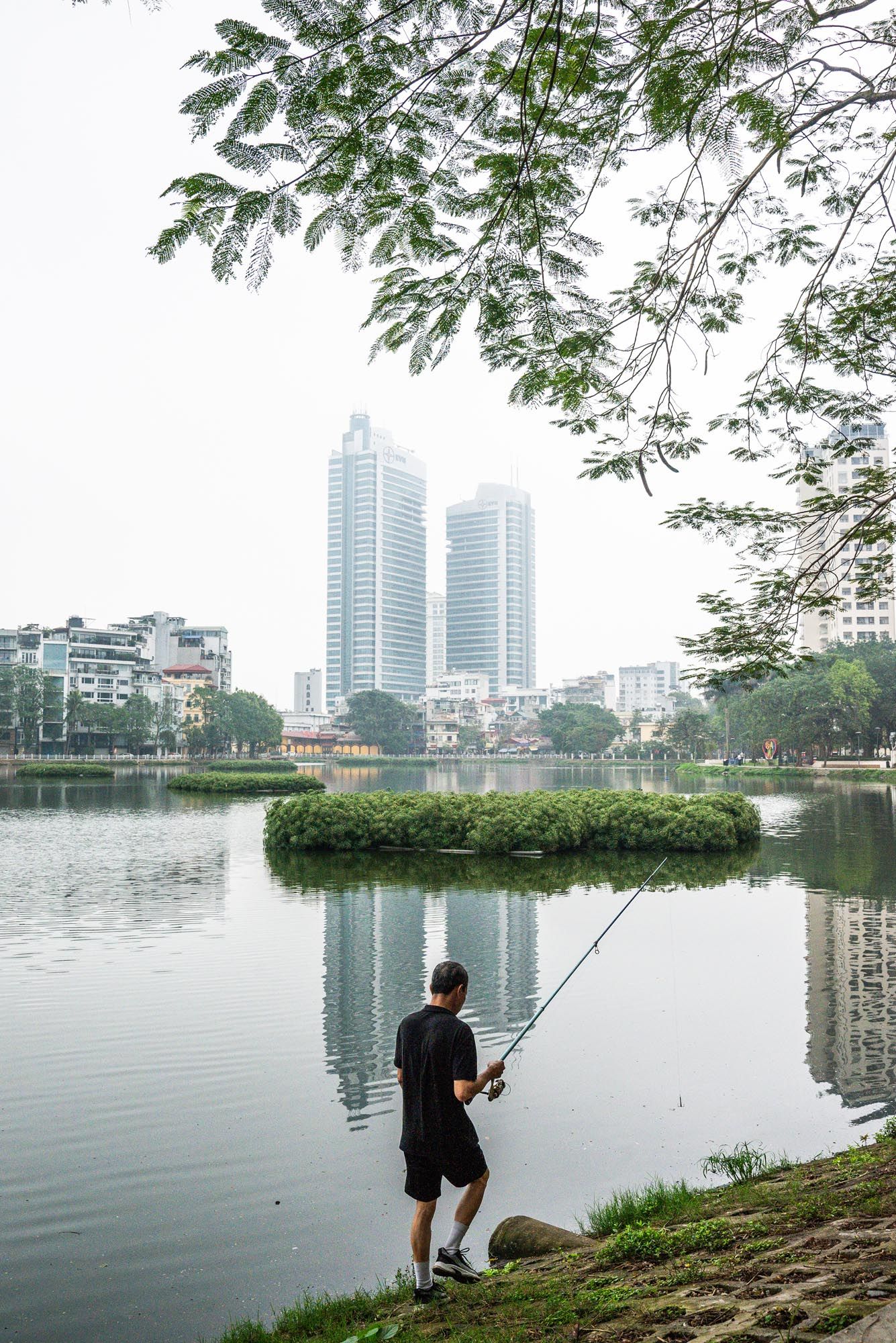 A person fishes by a lake with modern skyscrapers and lush trees in the background, creating an urban oasis scene.