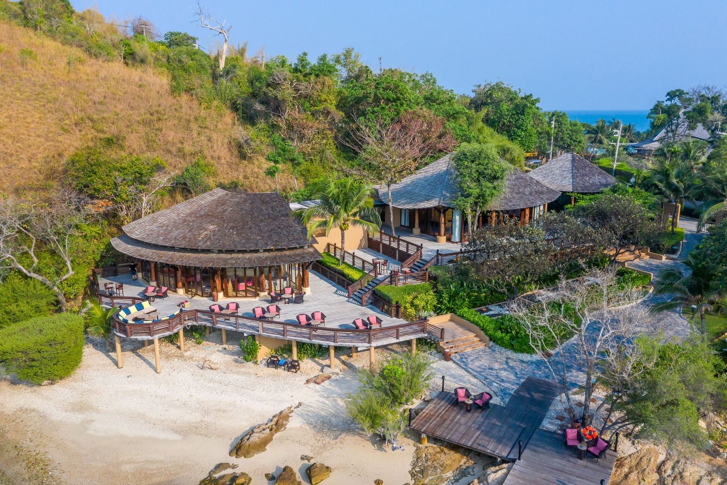 Aerial view of a tropical beach resort with thatched-roof buildings and outdoor dining areas overlooking the ocean.