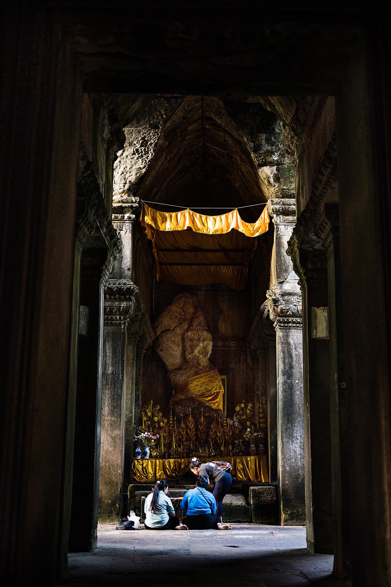 Tourists sit in prayer at a dimly lit Buddhist temple shrine with ornate stone columns and golden decorations.