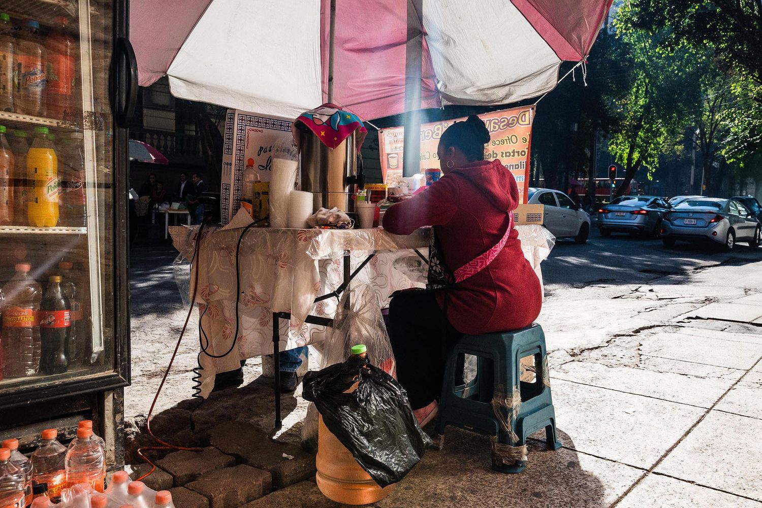 A street vendor prepares food at an outdoor cart with pink umbrella while customers wait on the sidewalk.