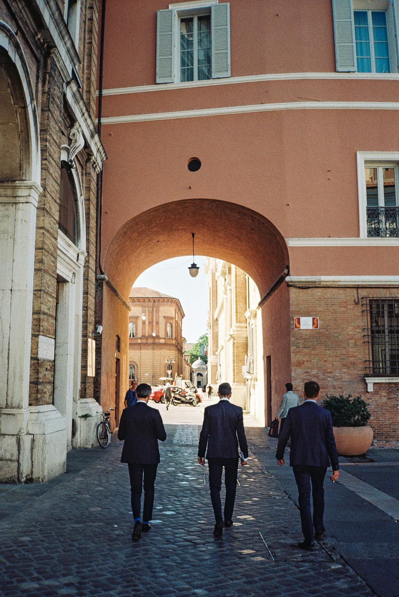 A sequence of people walking through an arched passageway with peach-colored walls and cobblestone flooring.