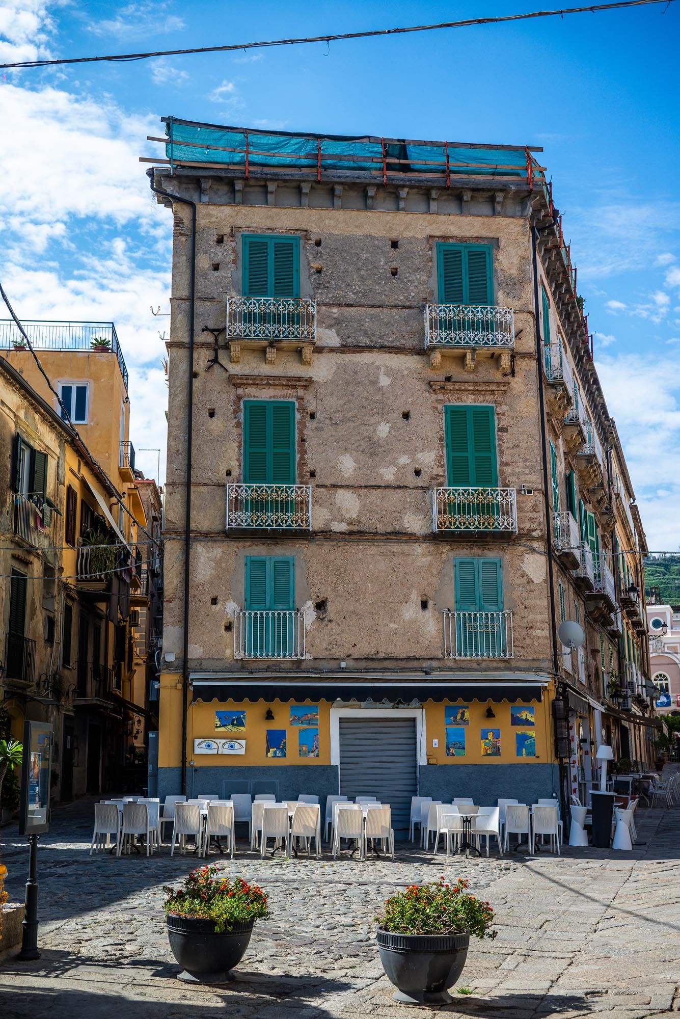 Historic Italian building with turquoise shutters and ornate balconies stands on a corner in Sicily with outdoor cafe seating.