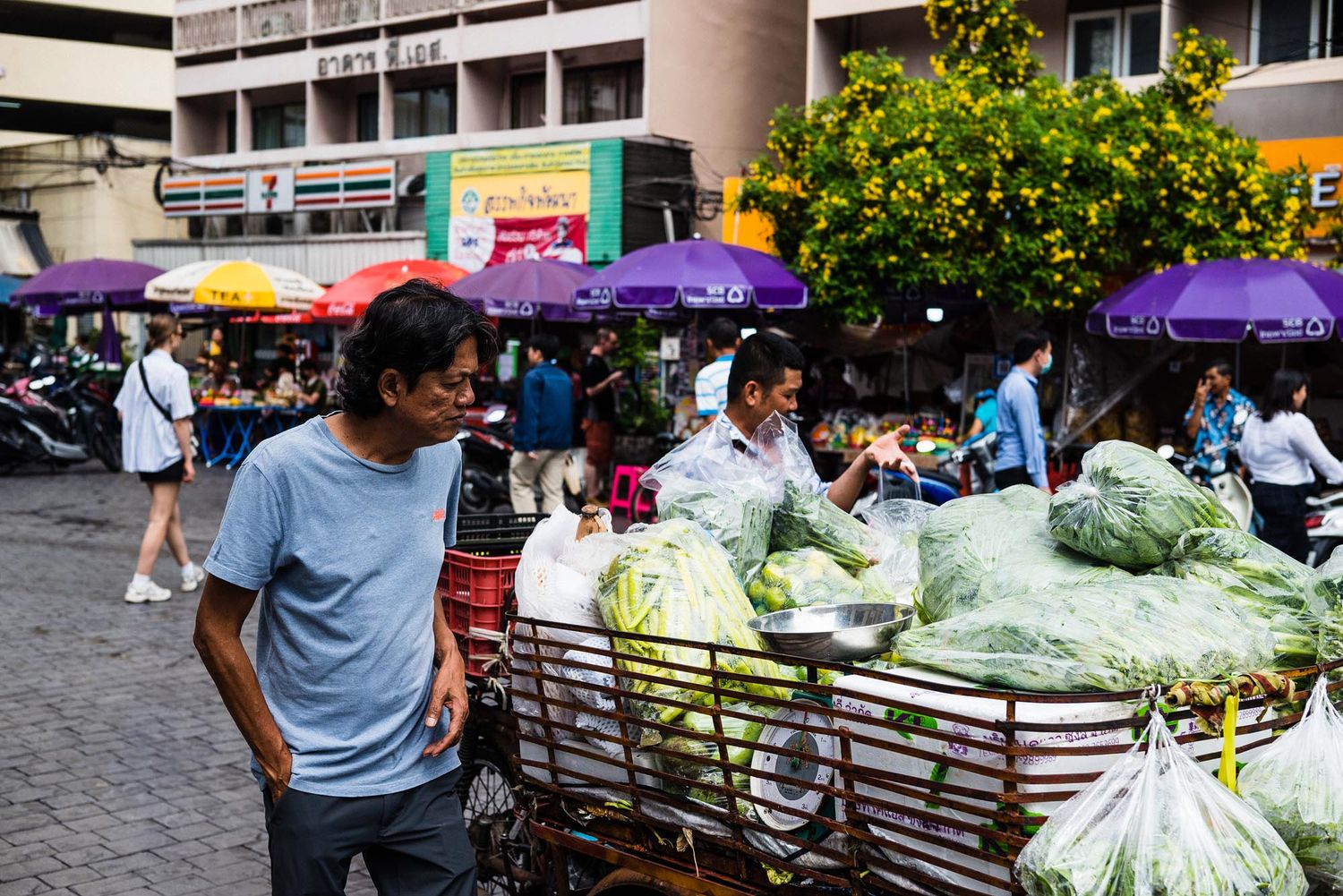 Colorful market stalls with purple umbrellas and fresh produce displayed under a sunny sky at an outdoor farmers market.