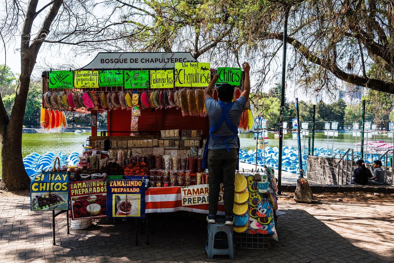 Colorful beach market stalls with vendors selling snacks and drinks under shady trees near the water.