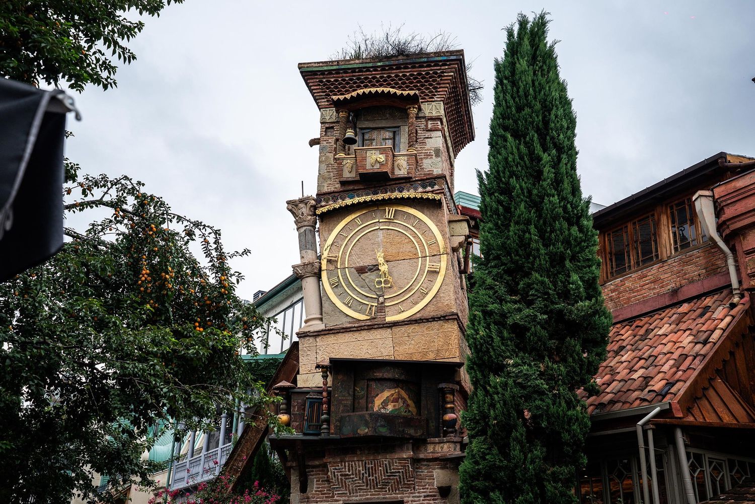 Historic clock tower with ornate architectural details rises between cypress trees and red-tiled roofs in Tbilisi, Georgia.