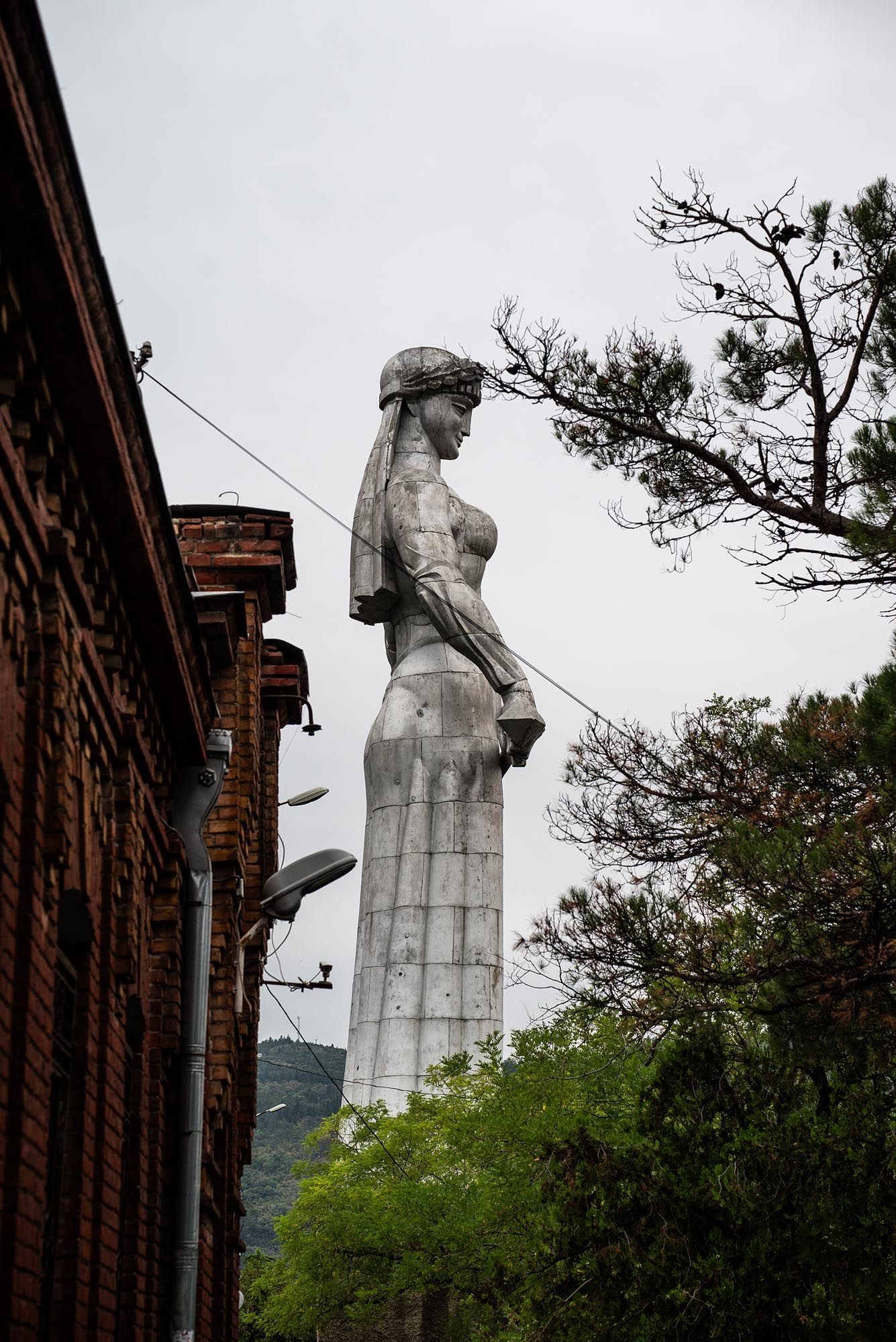 Monumental stone statues stand tall against a cloudy sky, surrounded by trees and brick buildings in the background.