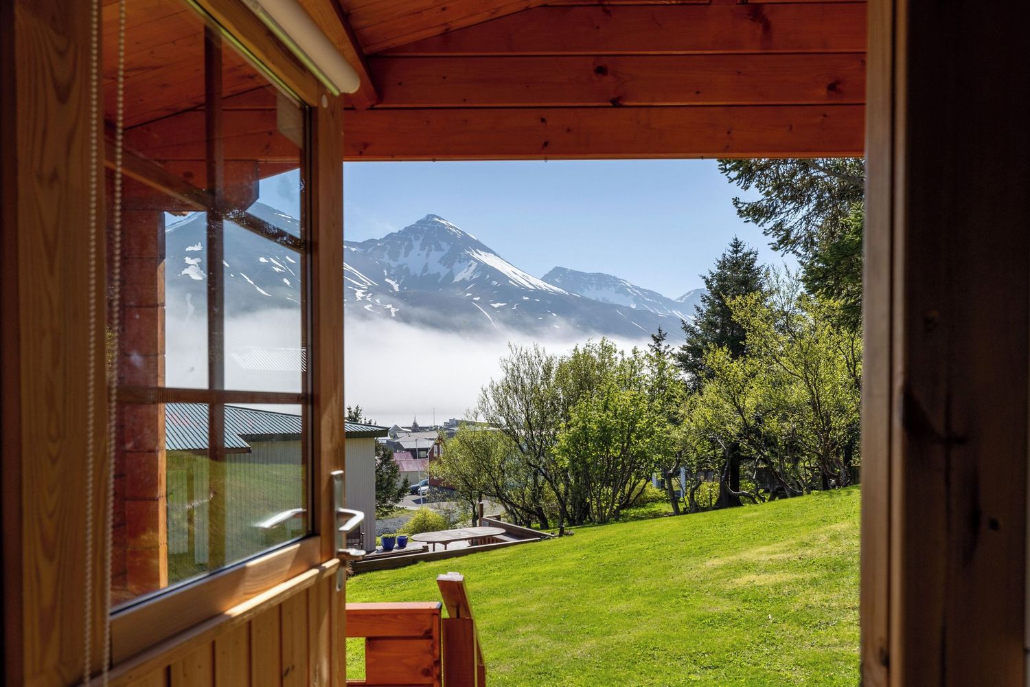 Panoramic mountain view through a wooden window frame with snow-capped peaks and green meadows in the distance.