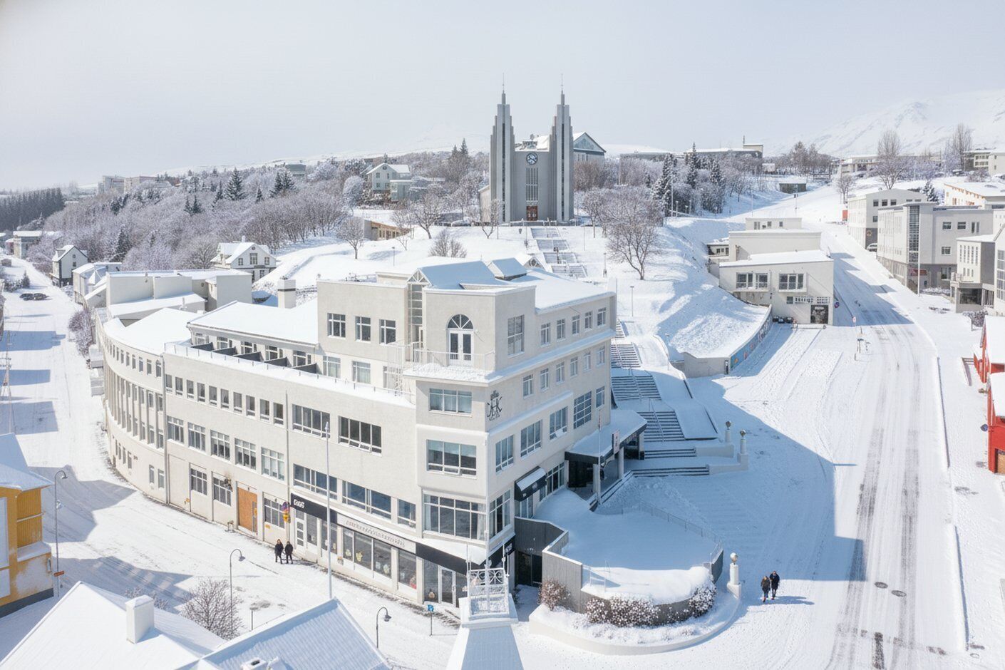 Aerial view of snow-covered art deco buildings at a winter resort with ski slopes and spires in the background.