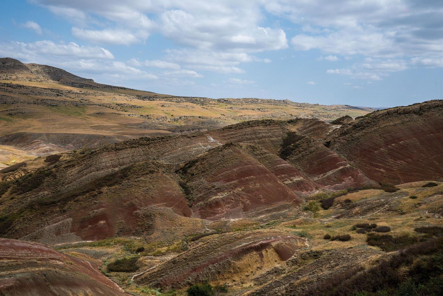 Winding dirt roads traverse through reddish-brown hills and valleys under a blue sky with scattered clouds.