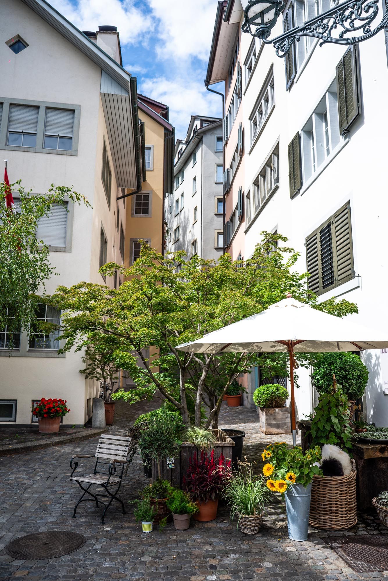 Charming European courtyard with cobblestone pavement, potted plants, outdoor furniture, and white umbrellas between buildings.