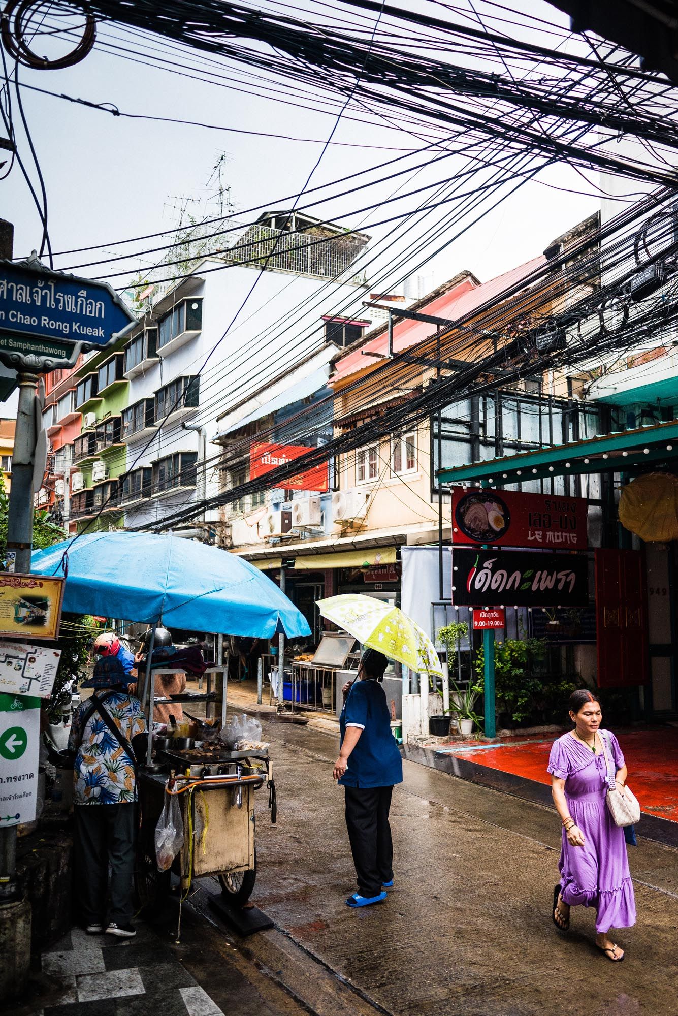 A rainy street scene in Bangkok with umbrellas, tangled power lines, and street vendors along the busy urban sidewalk.