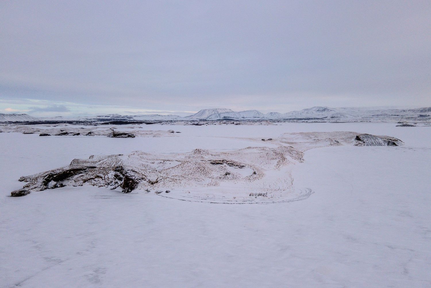 A snow-covered winter landscape with distant mountains under an overcast sky and curving frozen terrain.