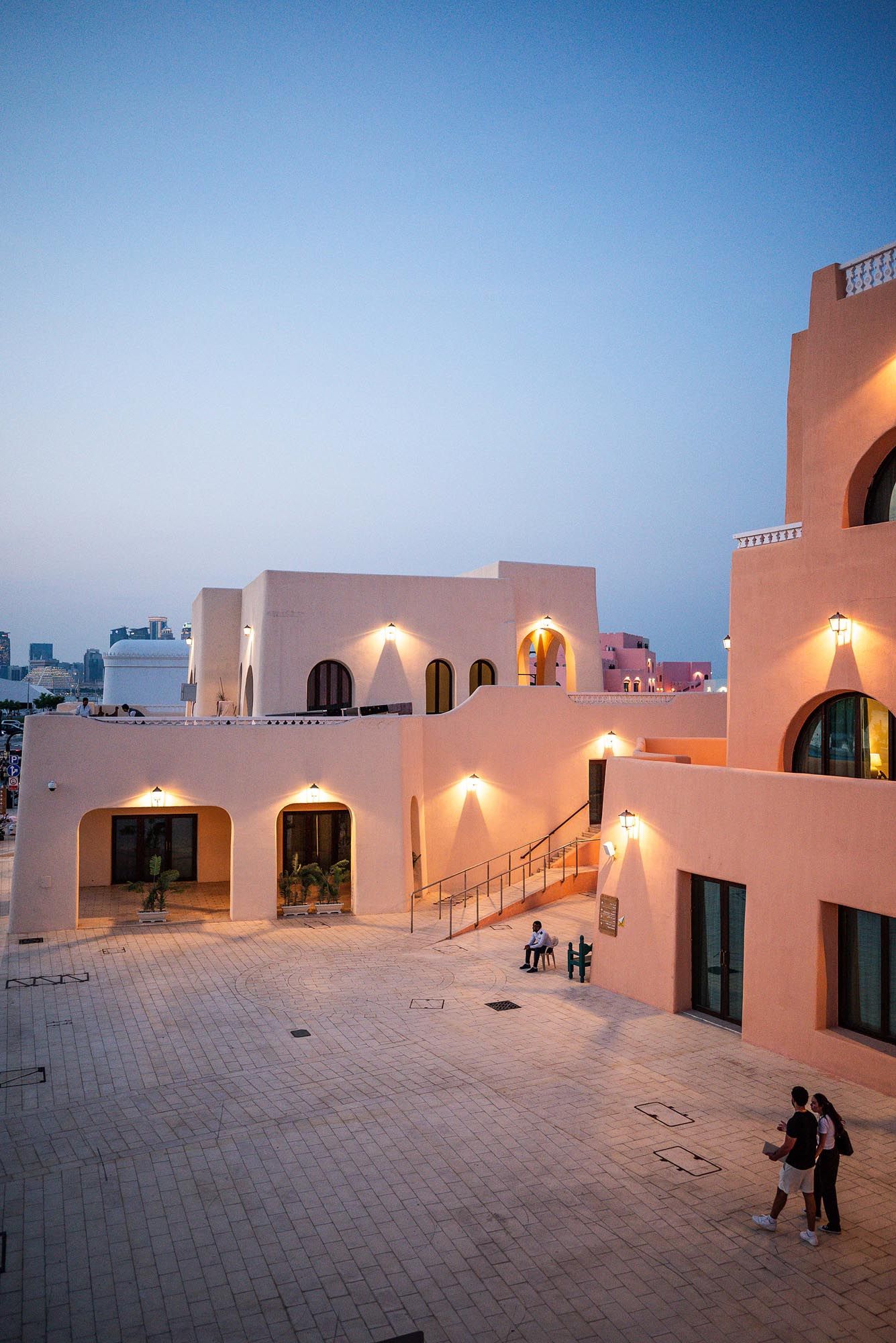 People walking on a modern white rooftop terrace with glowing lights at dusk against a blue sky.
