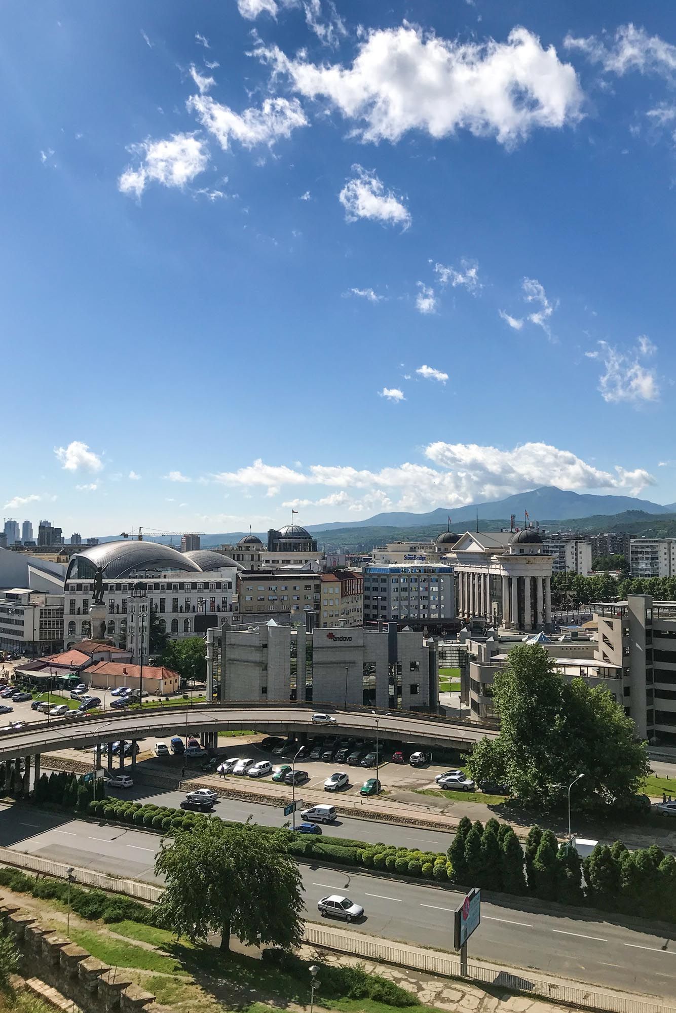 Panoramic cityscape view of an urban area with buildings, parking lots and mountains in the background under blue sky.