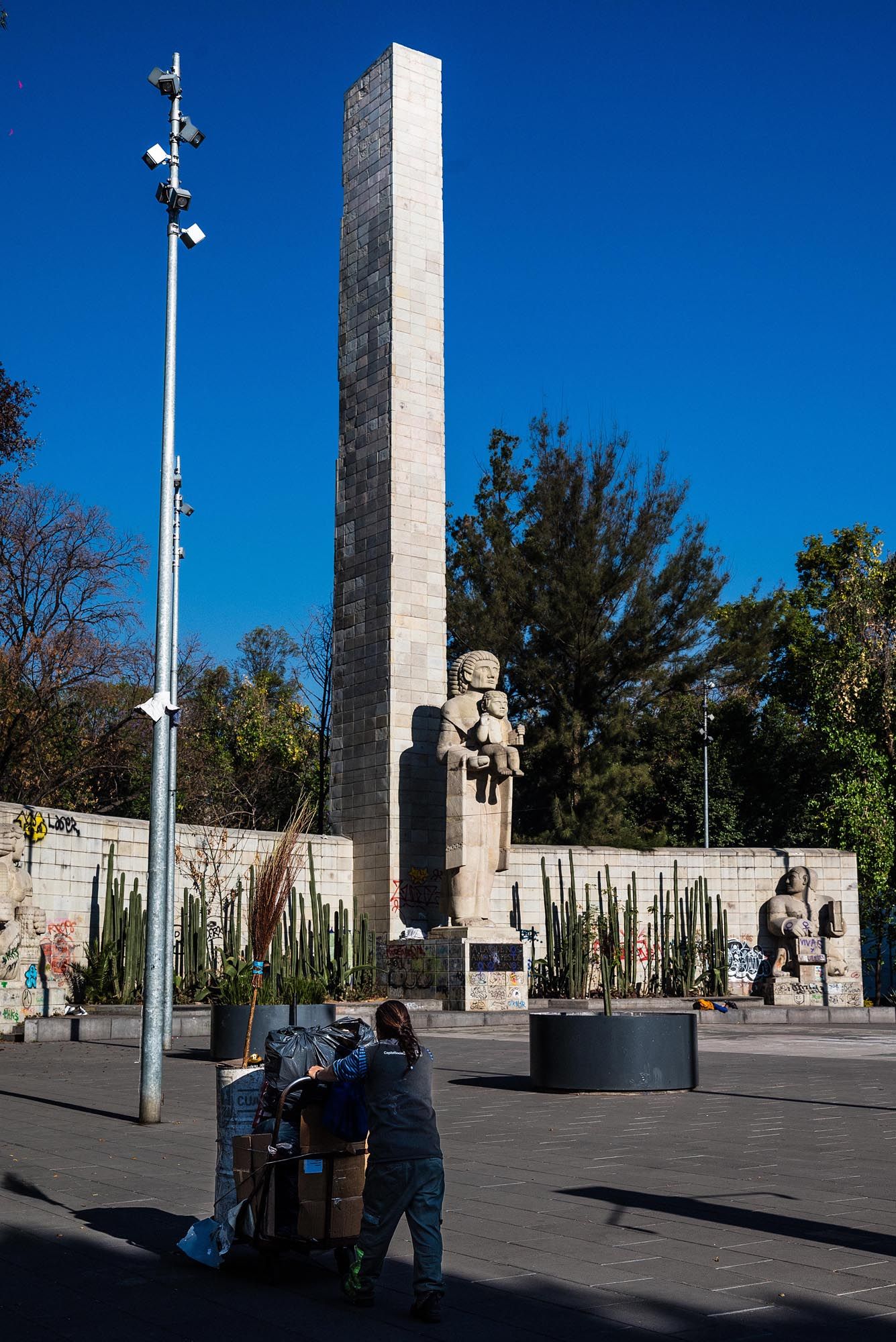 Several tall stone pillars stand against a bright blue sky in a public plaza with people walking nearby.