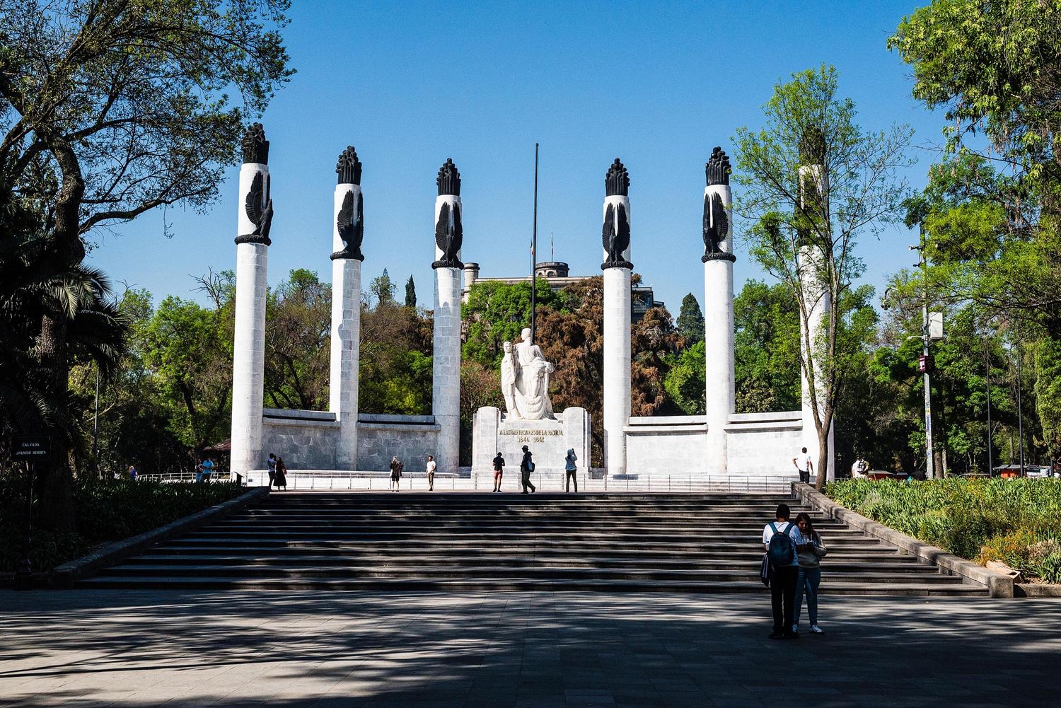 A panoramic view of the Heroes Monument in Mexico City featuring white pillars and black sculptures against a blue sky.