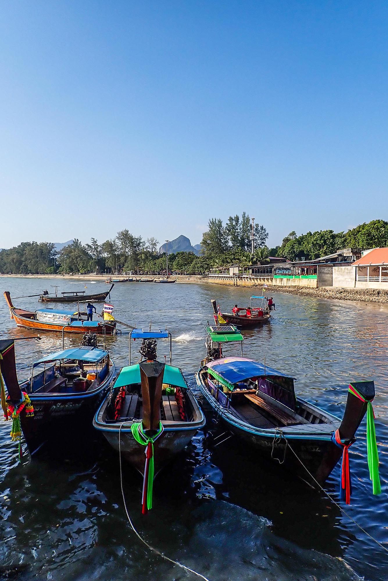 Traditional Thai longtail boats moored along the shoreline at sunset with colorful ribbons tied to their bows.