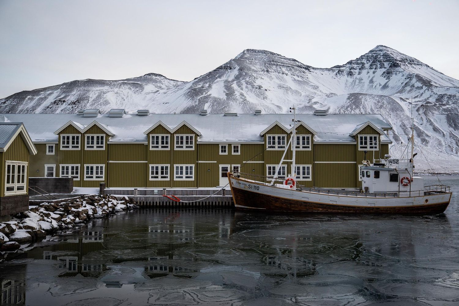 Row of wooden residential buildings along a snowy mountain coastline in a remote Arctic settlement.