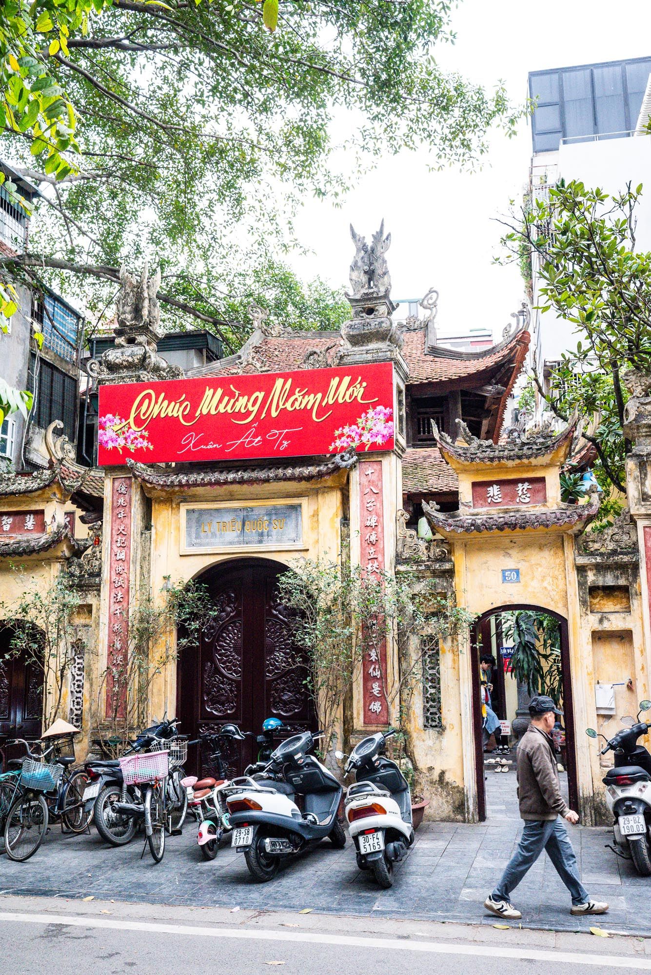 Scooters parked outside traditional Vietnamese temple entrance with red banner and ornate architecture in Hanoi, Vietnam.