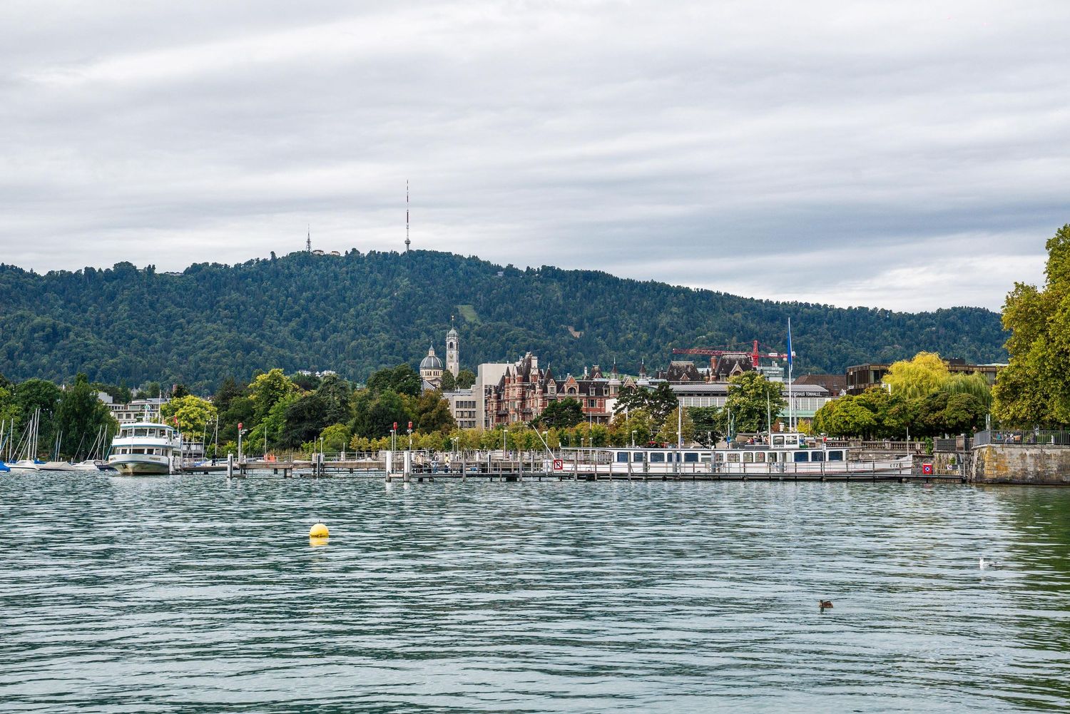 A panoramic view of a marina with boats docked along the waterfront against a backdrop of forested hills and cloudy skies.