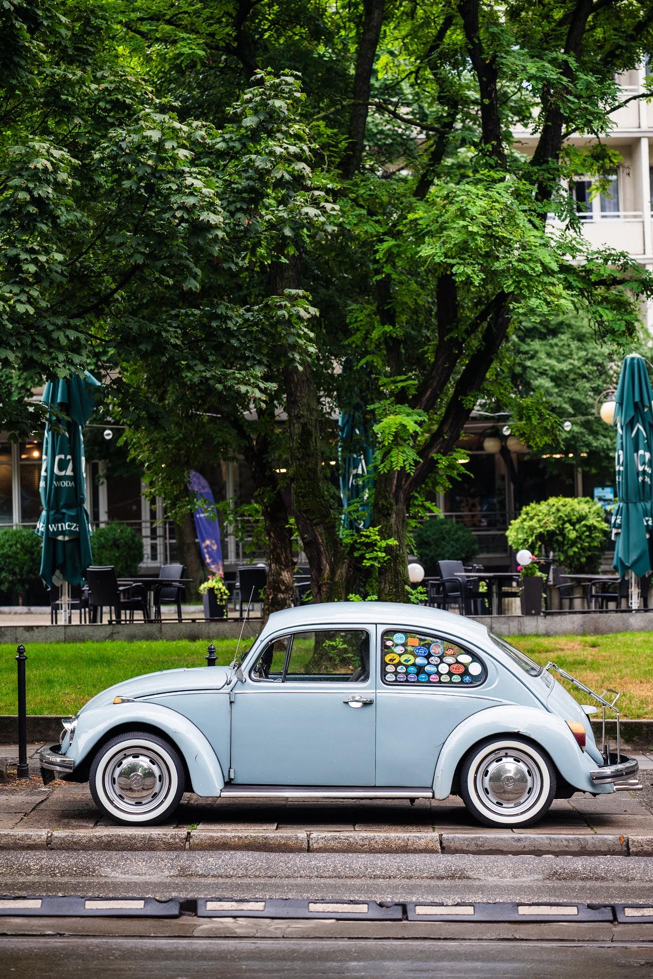 A classic light blue Volkswagen Beetle parked on a street with lush green trees and outdoor cafe seating in the background.