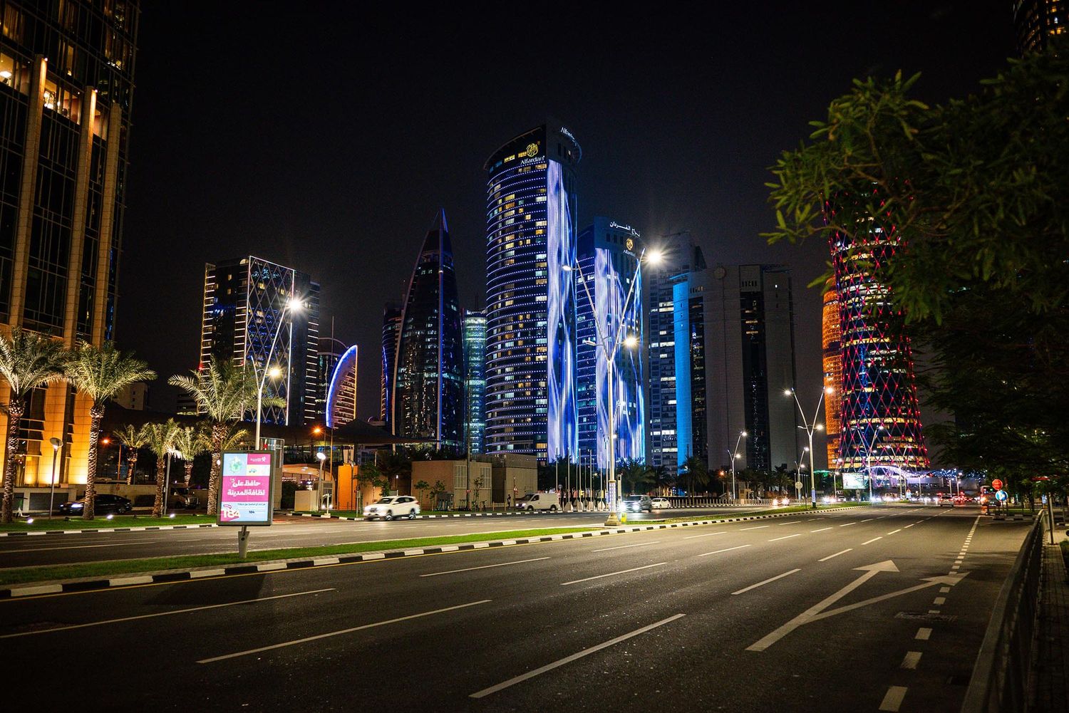Illuminated city skyline at night with modern skyscrapers and empty streets reflecting colorful lights.