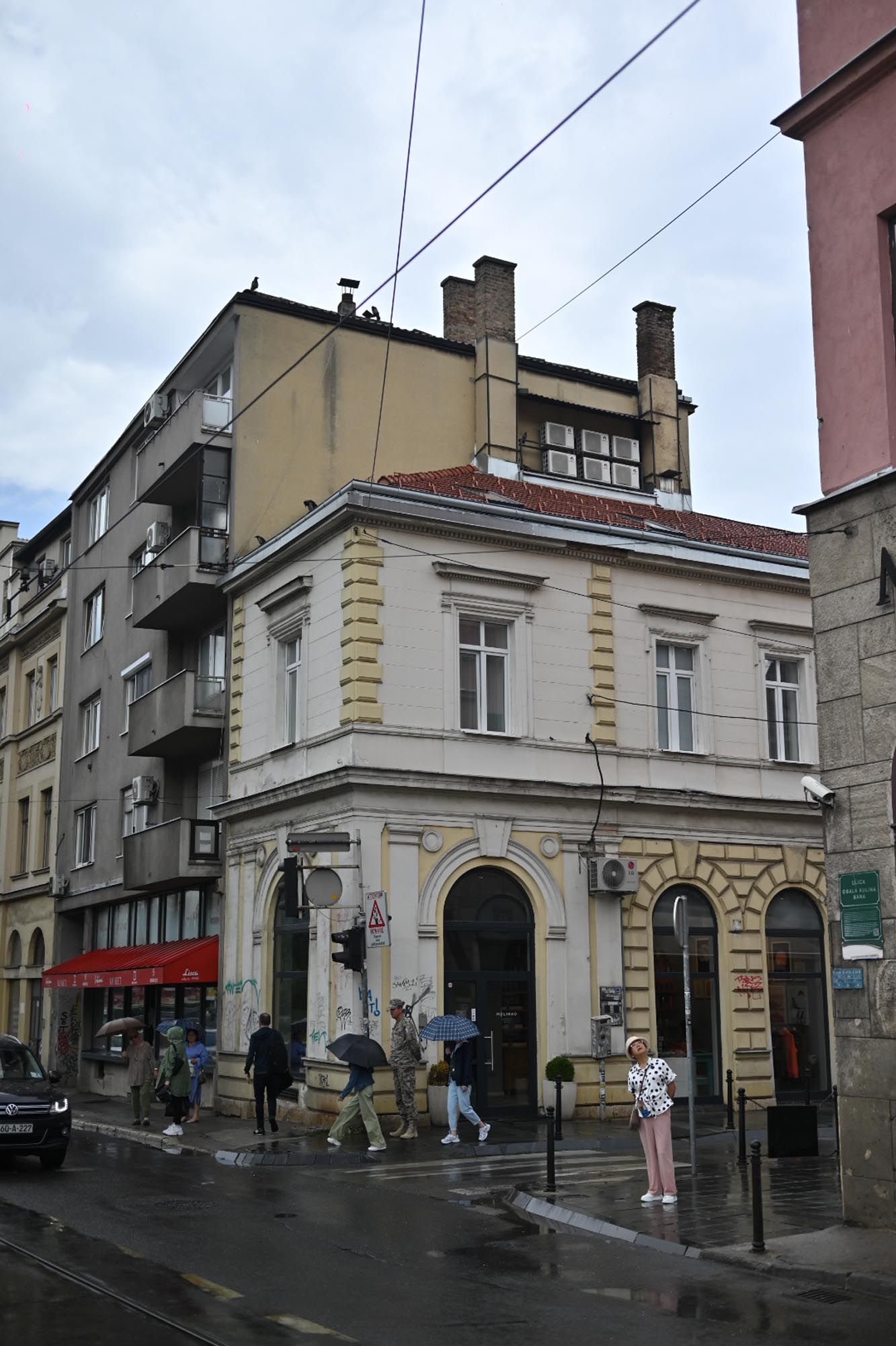Historic corner building with ornate yellow facade and arched entrances on a rainy street in an urban European setting.