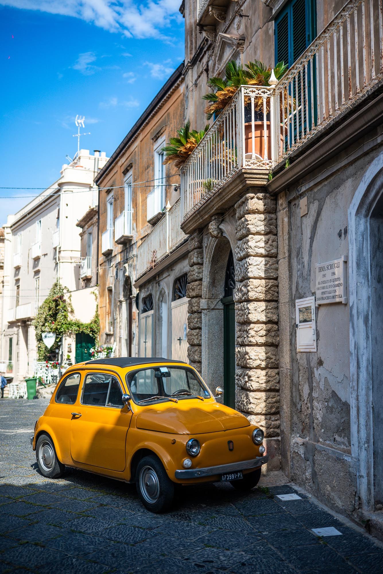 A bright yellow vintage Fiat 500 parked on a narrow cobblestone street in an old Italian town with historic buildings.