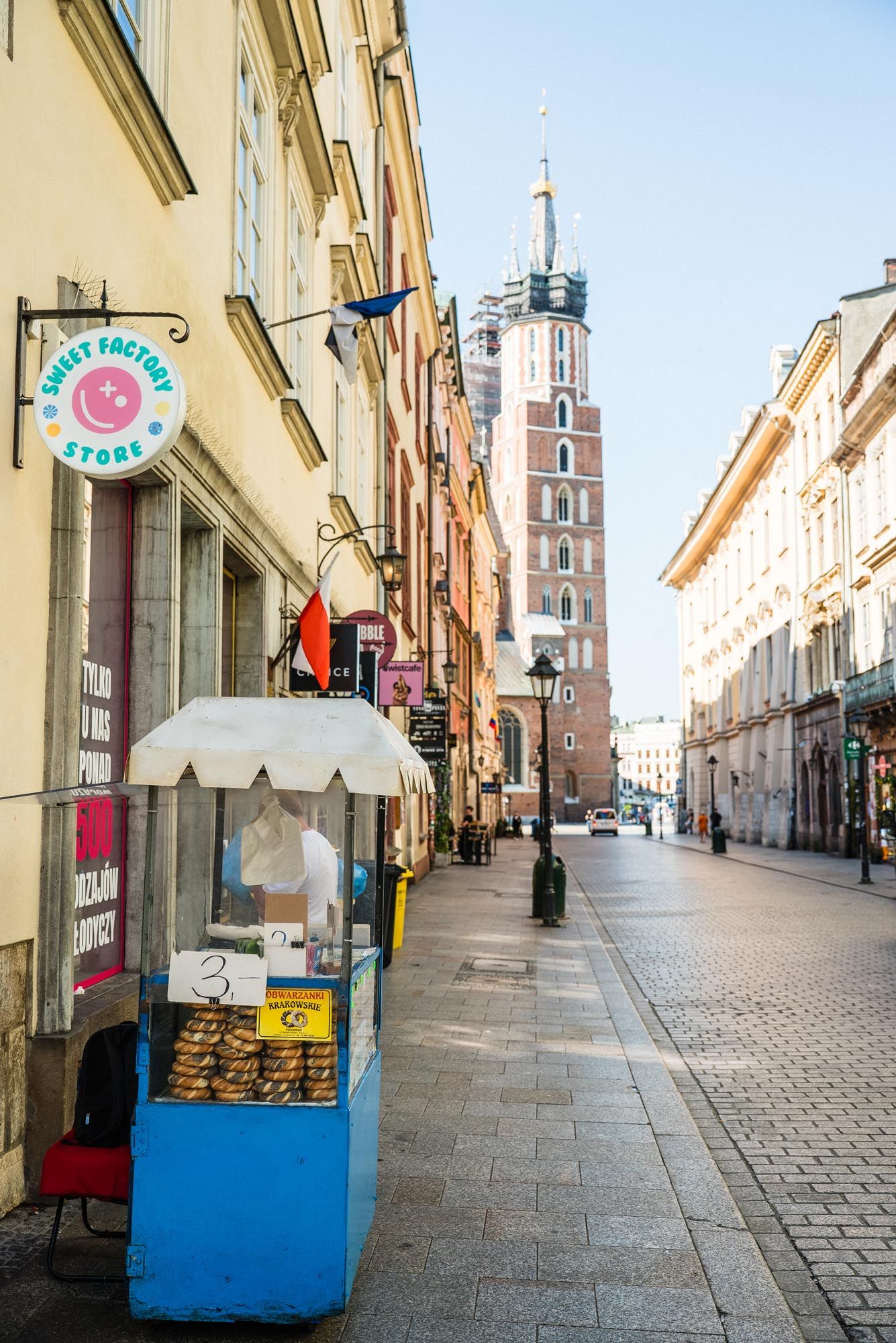 A cobblestone street in Krakow's Old Town with St. Mary's Basilica tower and a small blue street vendor kiosk.