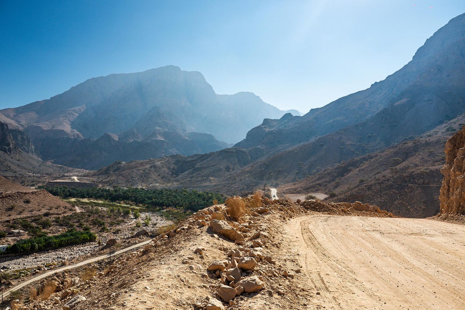 A winding dirt road curves through dramatic mountain terrain with rugged peaks and cliffs in the background. Wadi Al Arbeieen