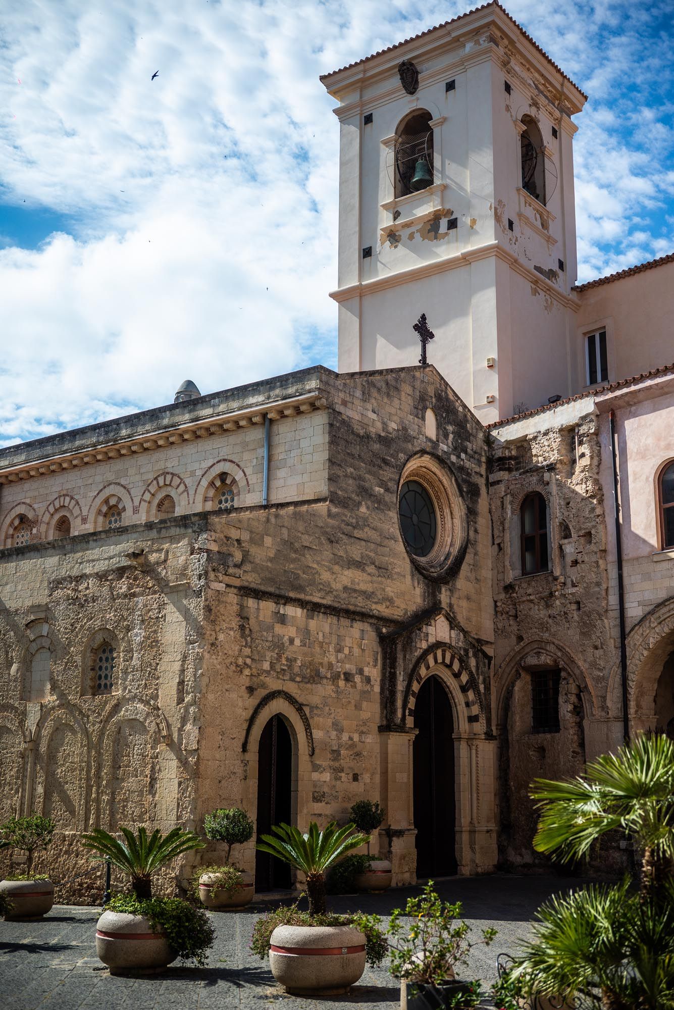 Ancient stone church courtyard with white bell tower and palm plants under blue cloudy sky in Mediterranean style.