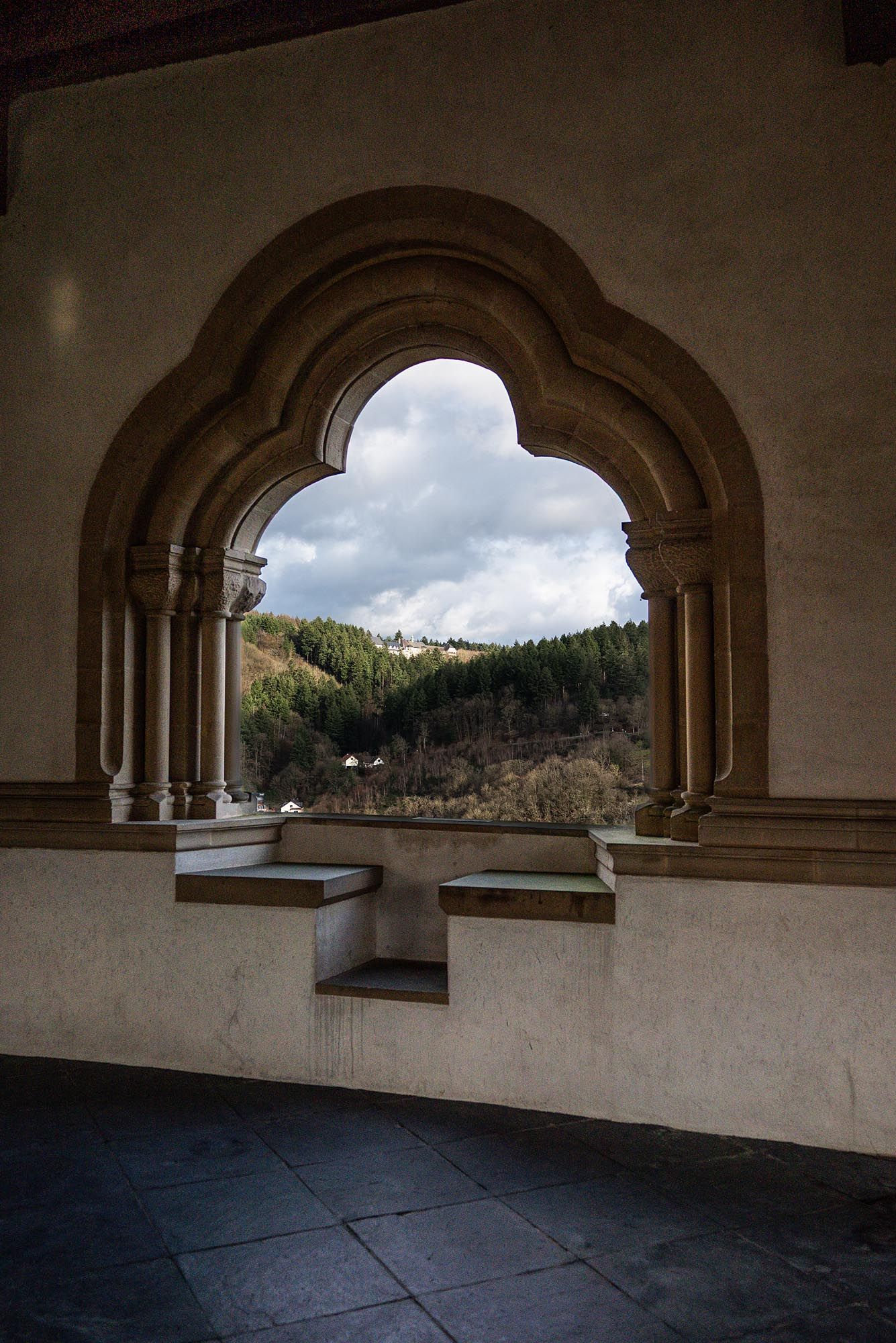 A series of arched stone windows in a monastery overlook a scenic mountain landscape on a cloudy day.