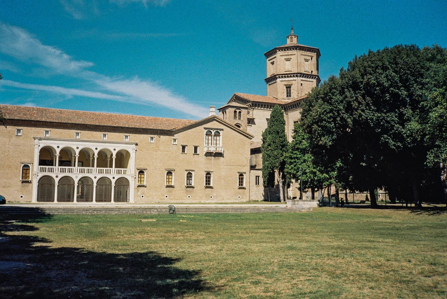 Historic stone monastery buildings with arched windows and columns stand against a bright blue sky on a grassy landscape.