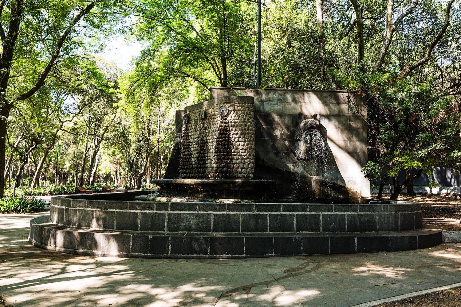 A scenic stone path winds through a lush green park with dappled sunlight filtering through the trees.