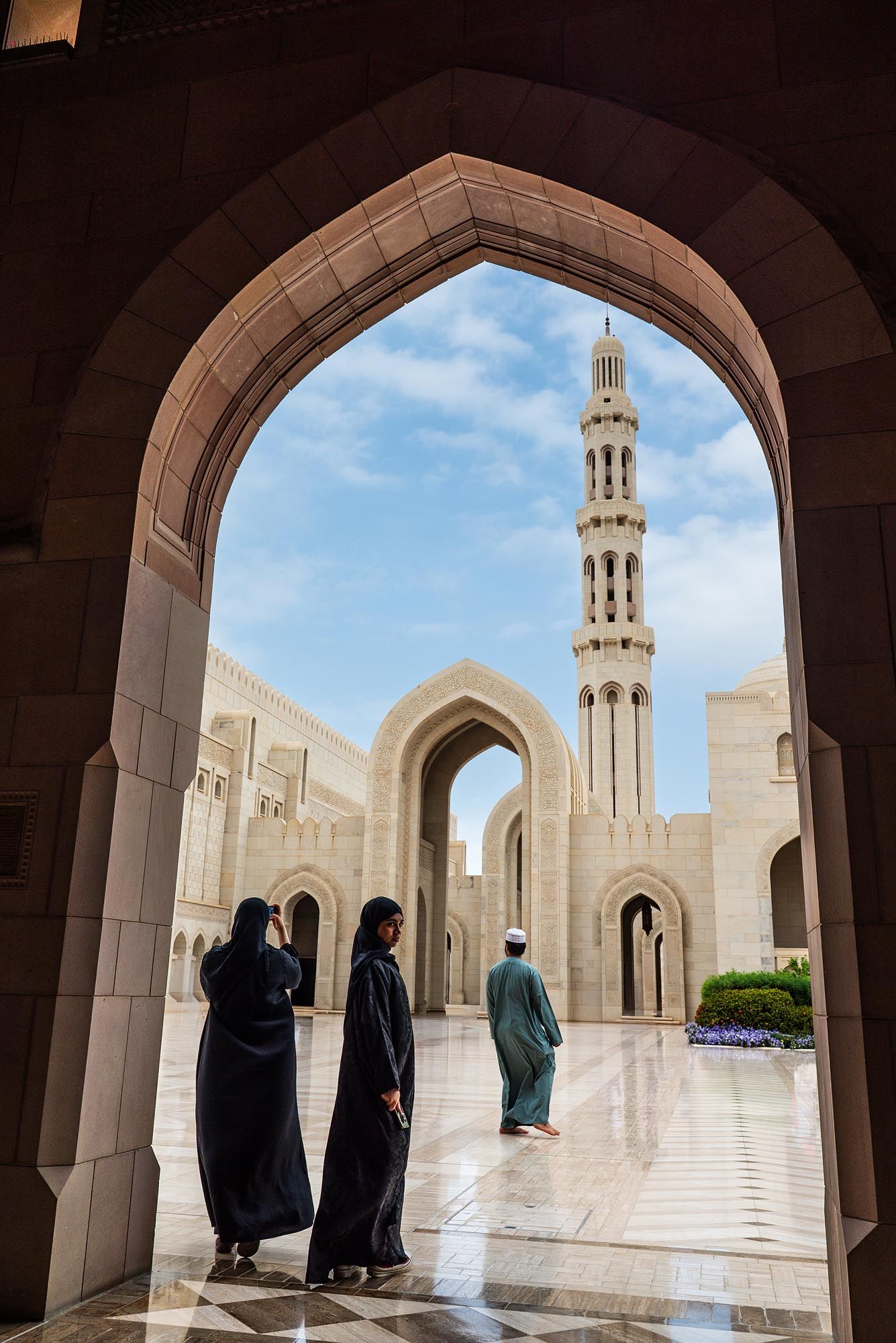 View through arched doorway of Sultan Qaboos Grand Mosque in Muscat, Oman with visitors in traditional dress.