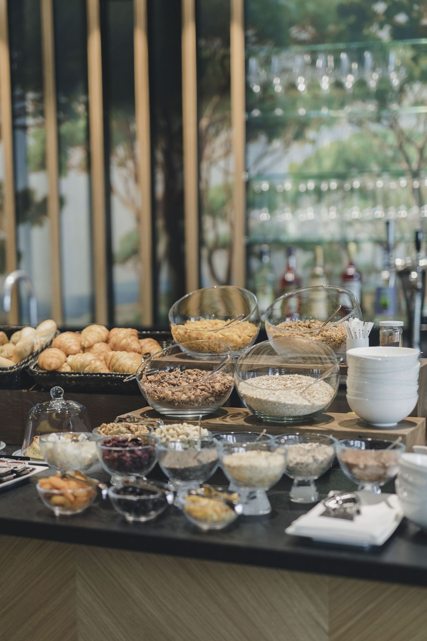 A rustic breakfast buffet table displays various cereals, nuts, dried fruits and granola in glass bowls with string lights above.
