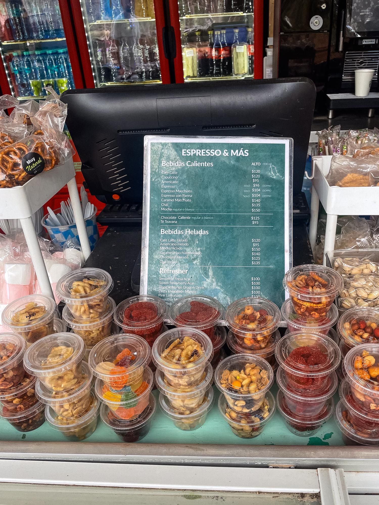 Clear plastic containers filled with various snacks and nuts displayed in front of a menu board at a coffee shop.