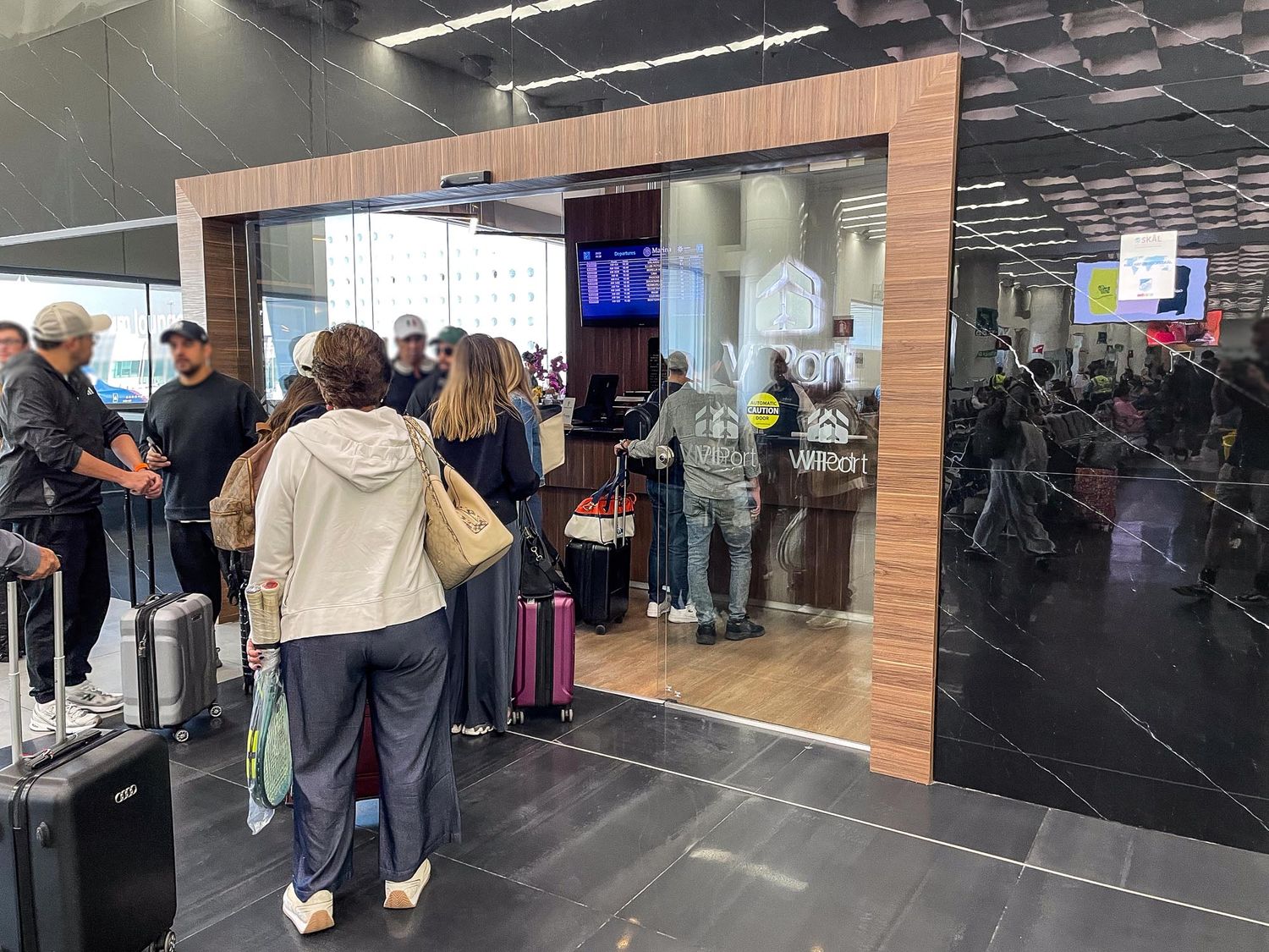 Passengers wait in line at an airport terminal with luggage and flight information displays visible in the background.