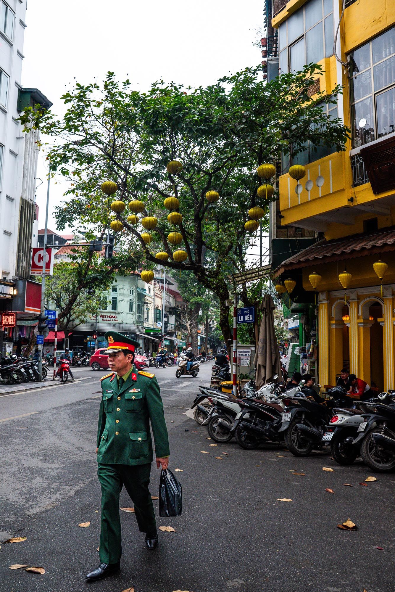 Busy street in Vietnam with yellow lanterns hanging above, parked motorcycles and a tree providing shade.