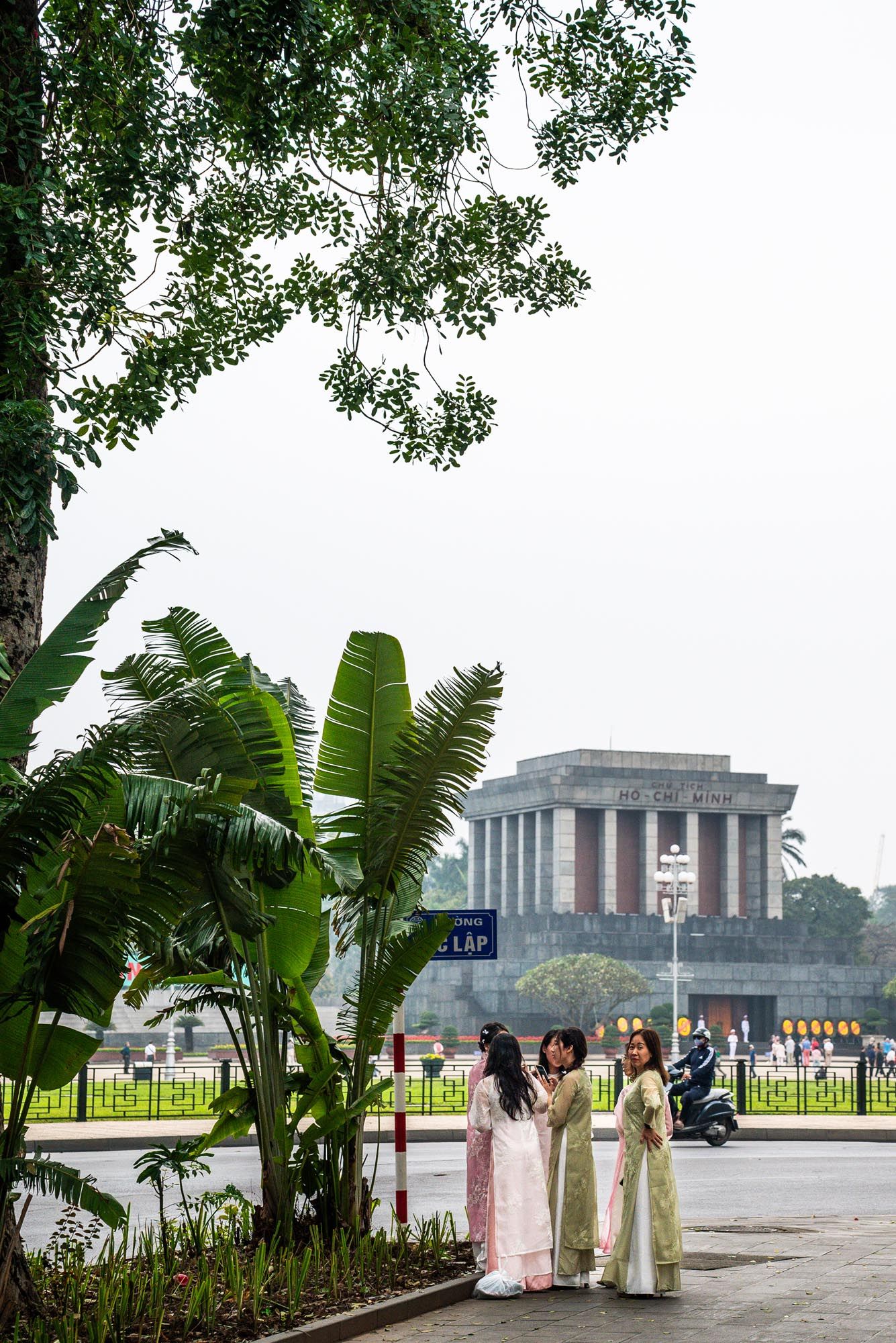 People walking near tropical palm trees with a modern building complex visible in the background in Hanoi, Vietnam.