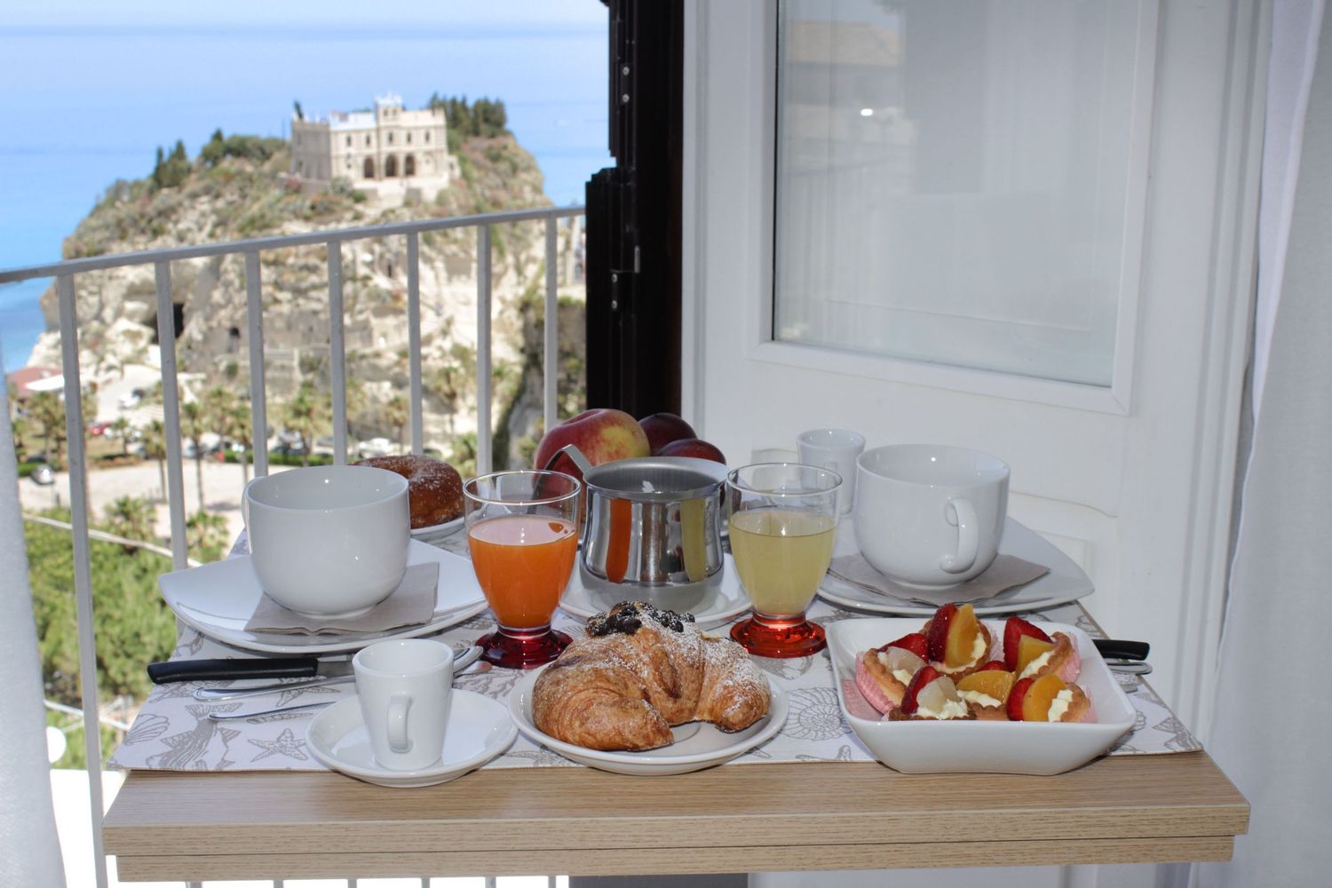 Breakfast spread on a white table overlooking a Mediterranean coastal town with hillside buildings and blue sea views.