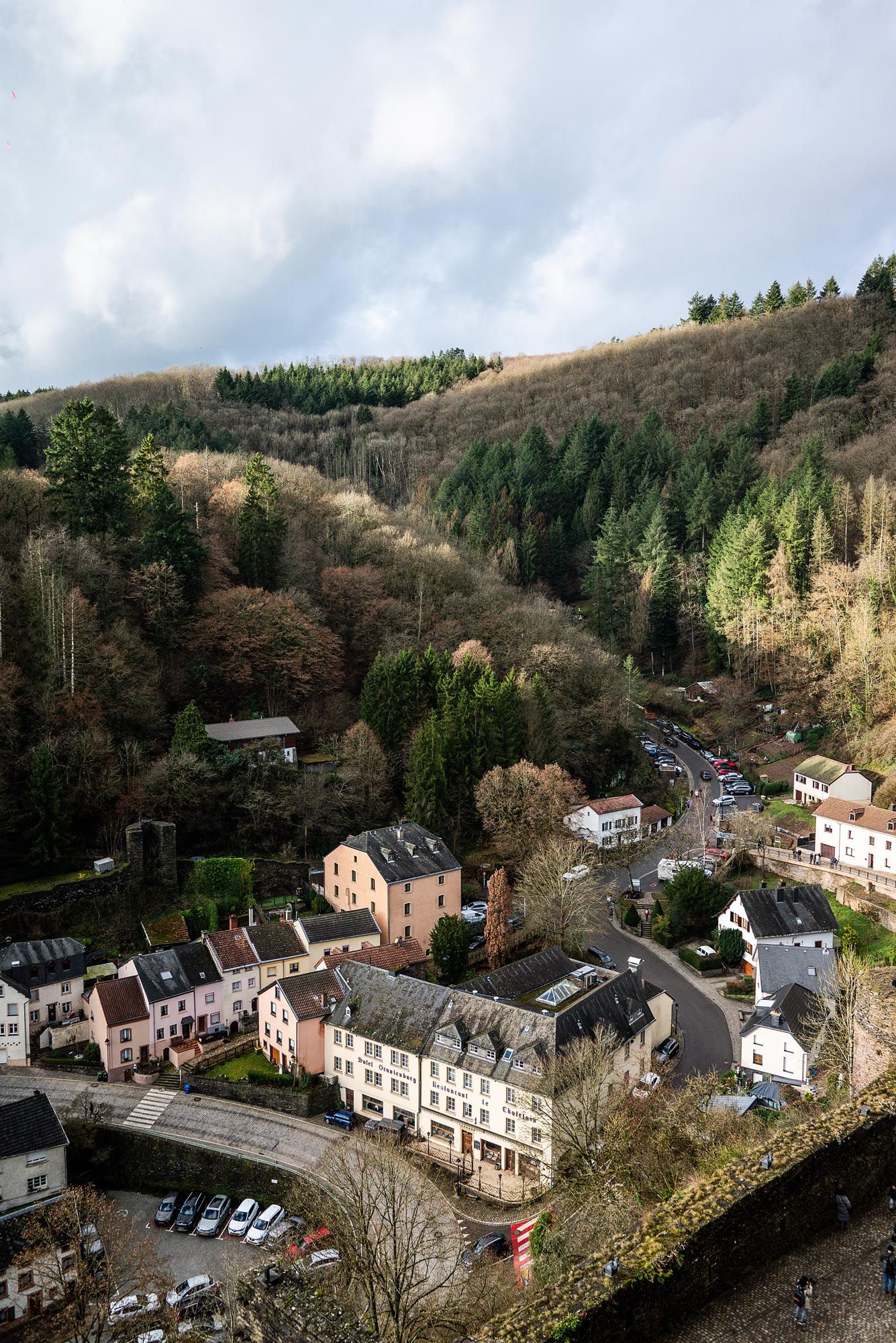 Aerial panoramic view of a quaint village nestled in a forested valley with traditional houses and parked cars.