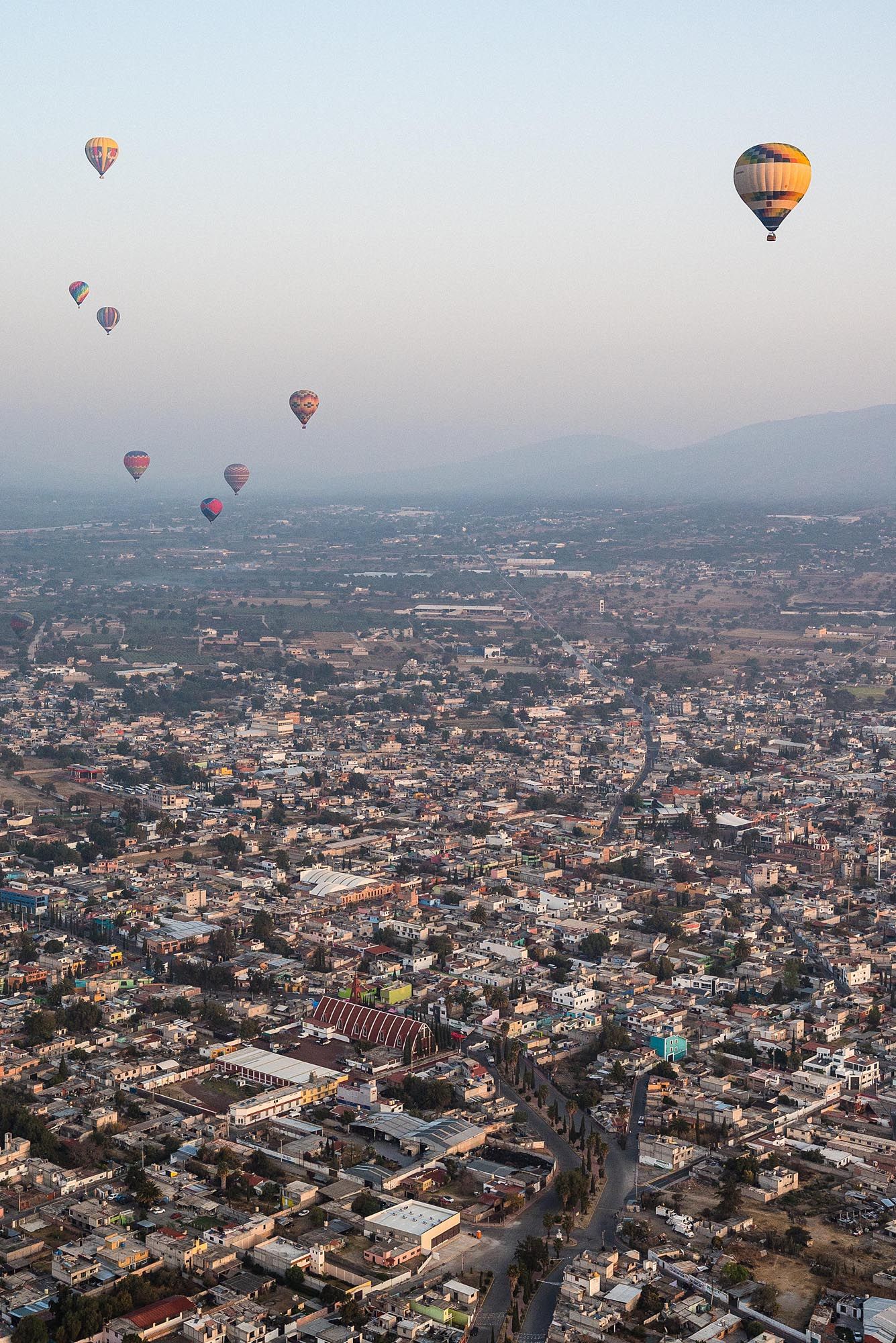 Hot air balloons float above a sprawling Mexican cityscape at sunrise with mountains in the distance.