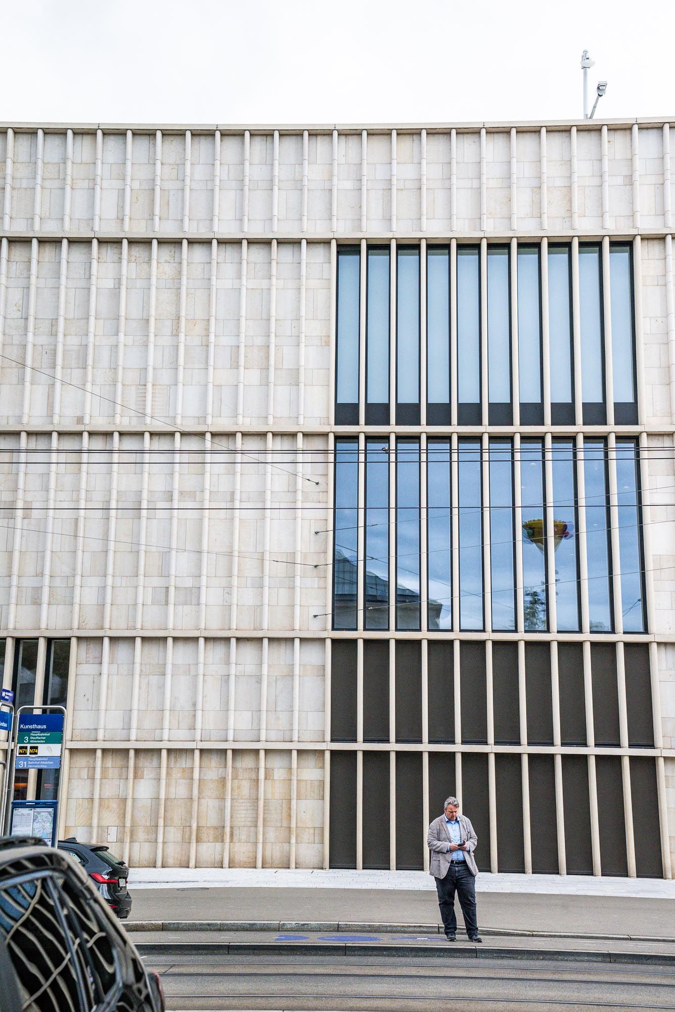 A person stands in front of a modernist concrete building facade with vertical lines and blue glass windows.