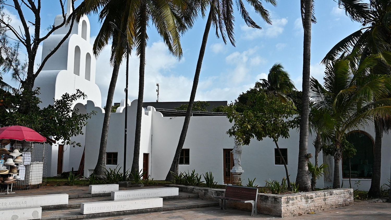 Palm trees line a pathway leading to white Mediterranean-style buildings under a bright blue sky.