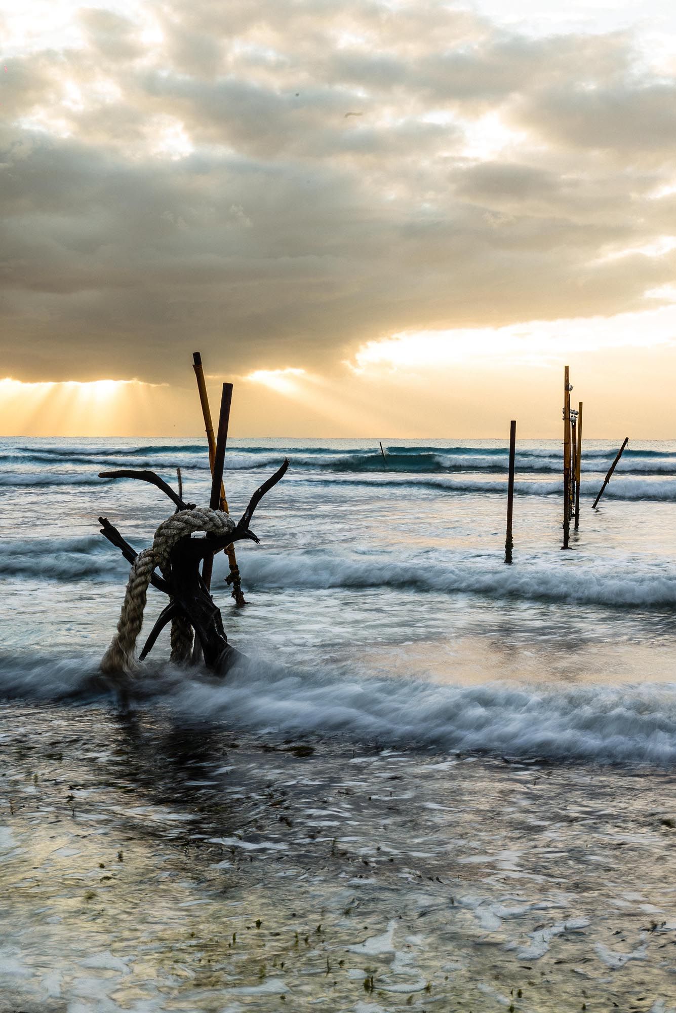 Dramatic sunset over ocean waves with wooden stilt poles silhouetted against golden cloudy sky at beach.
