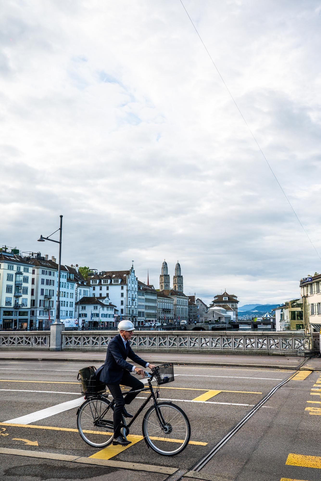 A cyclist rides past historic buildings along a waterfront promenade in Zurich on a cloudy day.