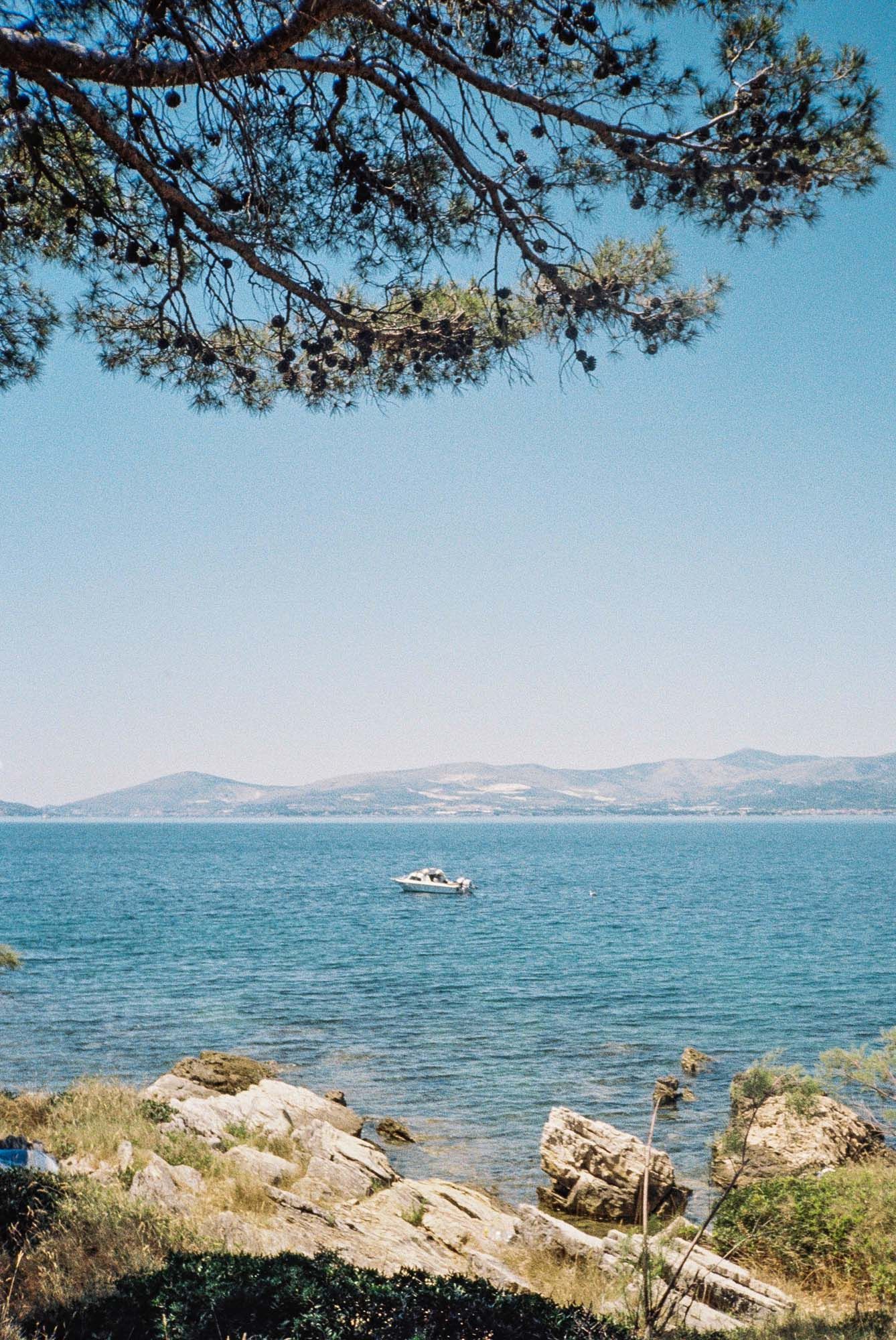 Panoramic Mediterranean coastal landscape with blue waters, rocky shore, pine trees, and distant mountains on a sunny day.